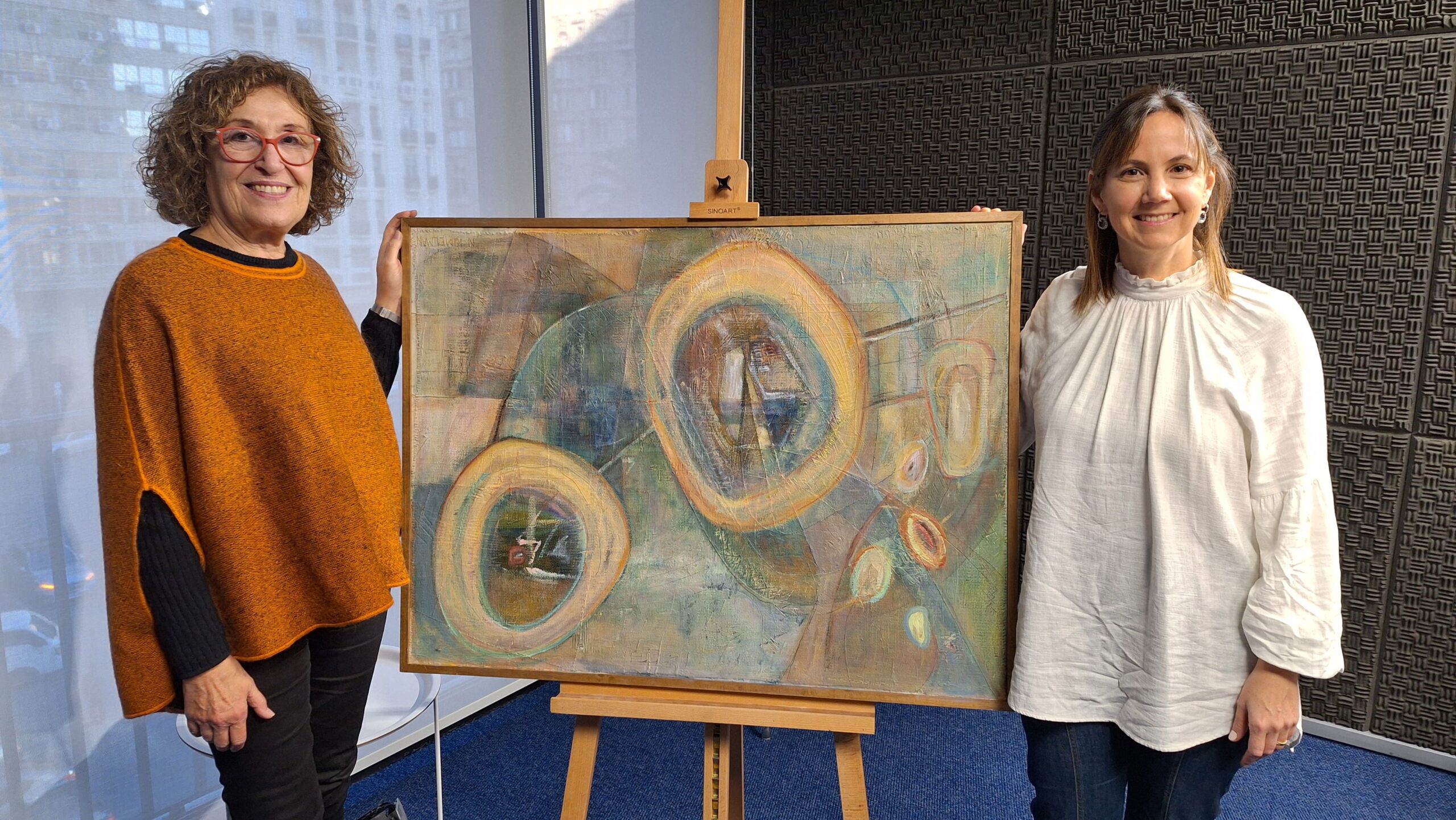 dos mujeres paradas entre un cuadro con tonos claros y figuras abstractas en un estudio de radio