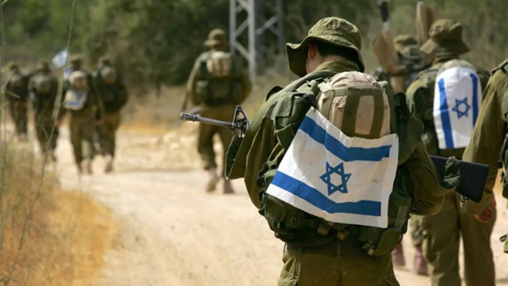 Militares israelíes caminando en camino de pedregullo con una bandera de Israel sobre sus mochilas